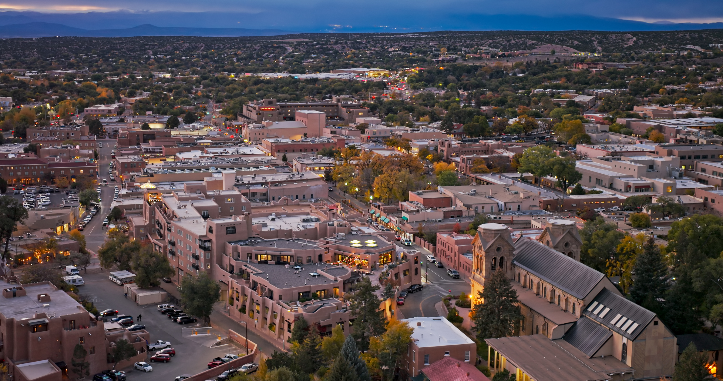 Aerial view of downtown Santa Fe's plaza area during blue hour, featuring the central plaza surrounded by adobe-style buildings and shops. The historic district displays characteristic southwestern architecture with flat-roofed structures in earth tones. Loretto Chapel with its Gothic Revival spire is visible on the right side of the frame. The plaza area serves as the city's cultural and commercial center, with tree-lined streets extending into surrounding residential neighborhoods. Parking areas and illuminated storefronts are visible throughout the district as evening lights begin to appear.